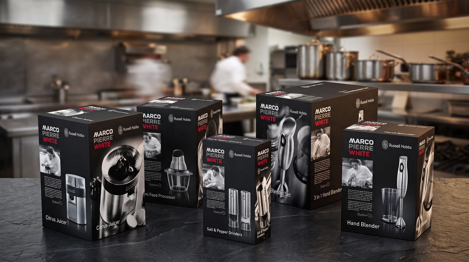 Marco Pierre White by Russell Hobbs packaging range: citrus juicer, mini food processor, salt and pepper grinders, 3 in 1 hand blender set and hand blender, on a dark worktop in a professional kitchen